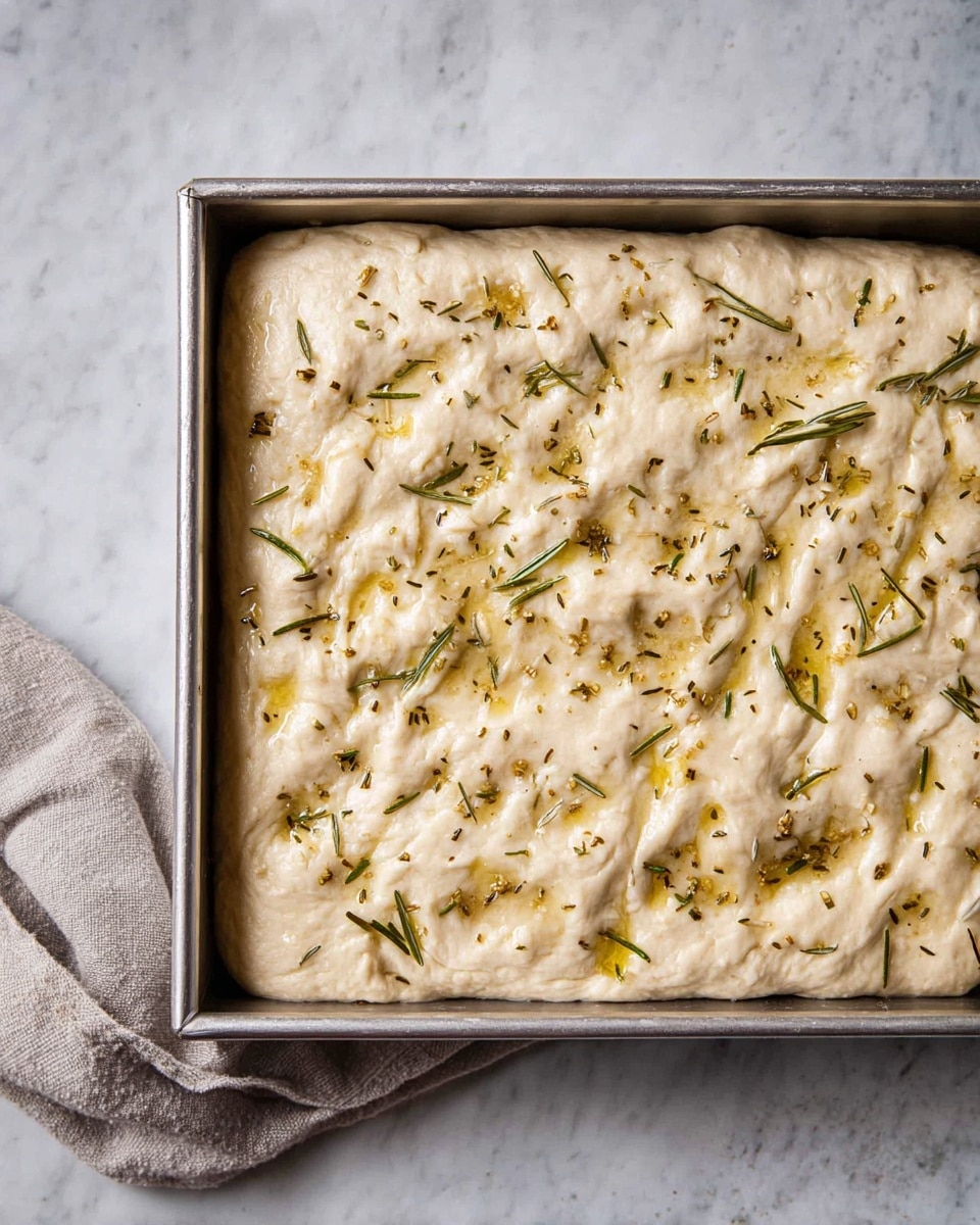 A rectangular metal tray holds one thick layer of pale, soft dough that fills the tray completely. The dough surface is uneven with small dimples and is sprinkled with small green herb pieces, likely rosemary and other dried herbs, along with some shiny spots of oil. The tray rests on a white marbled texture with a light gray cloth nearby. photo taken with an iphone --ar 4:5 --v 7