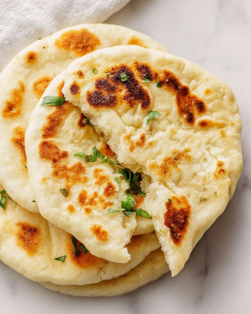 A stack of three round, soft flatbreads is shown on a white marbled surface, each flatbread having a golden brown spotted texture from cooking. The top flatbread is partially torn to reveal its fluffy, light interior, with some fresh green herb leaves peeking out from the bottom layer. The flatbreads have a pale, creamy color with uneven golden patches on their surface. photo taken with an iphone --ar 4:5 --v 7