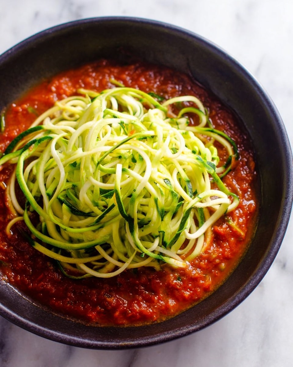 The image shows a black bowl filled with two layers: the bottom layer is a thick red tomato sauce with a slightly chunky texture, while the top layer is a loose pile of long, spiral-shaped zucchini noodles that are light green with some darker green strips from the skin. The noodles sit centered on the sauce, creating a strong color contrast between the red and green. The bowl is placed on a white marbled surface with soft natural light shining on it, highlighting the fresh and vibrant colors of the dish. photo taken with an iphone --ar 4:5 --v 7