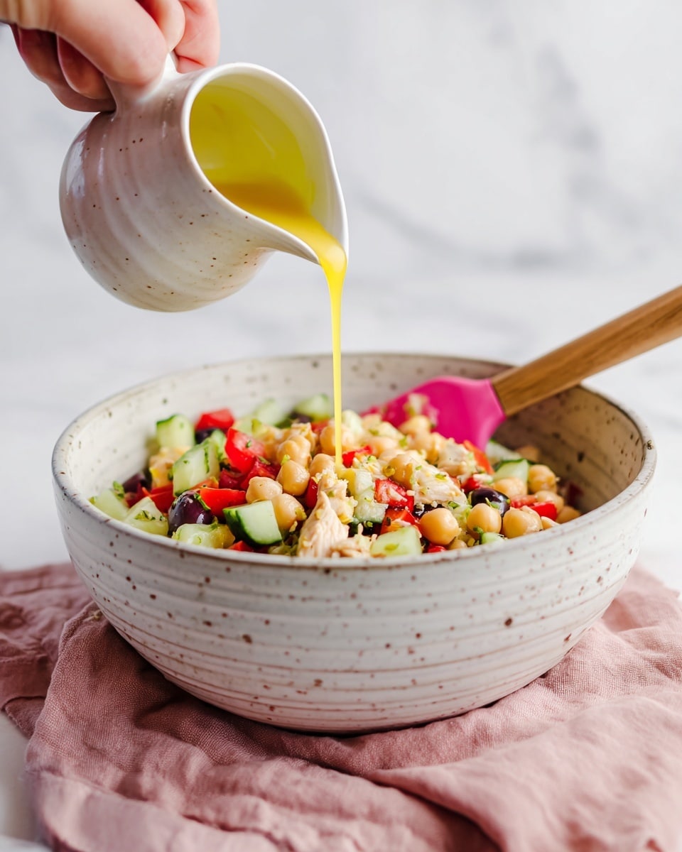 A white speckled bowl holds a colorful salad made of chopped cucumbers, chickpeas, olives, red bell peppers, and pieces of light brown chicken, mixed together and stirred with a spatula that has a pink silicone head and wooden handle resting inside the bowl. A woman's hand is pouring a bright yellow dressing from a small white ceramic jug into the salad. The bowl sits on a soft pink cloth on a white marbled surface, and the background is also white and blurred. photo taken with an iphone --ar 4:5 --v 7