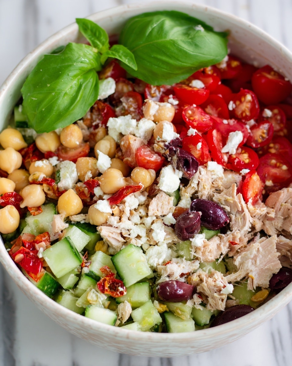 A colorful salad in a white bowl with a mix of peeled chickpeas, chopped cucumbers in green chunks, halved bright red cherry tomatoes, pieces of shredded light brown tuna, and chunks of white cheese scattered all over. There are also small pieces of dark purple olives and bits of red roasted pepper spread throughout. On top of the salad, there is a fresh green basil leaf as decoration. The bowl sits on a white marbled surface. photo taken with an iphone --ar 4:5 --v 7