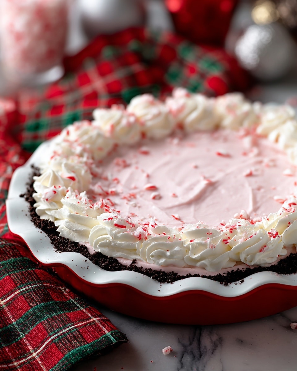 The image shows a pie with three clear layers placed in a white pie dish with a red scalloped edge. The bottom layer is a dark, crumbly crust, on top of which is a thick, smooth layer of pale pink cream. Around the edge of the pie, there is a ring of white whipped cream piped in soft swirls. Small crushed red and white peppermint pieces are scattered over the pink cream and the whipped cream, adding texture and color. The pie sits on a white marbled surface, with red, green, and white plaid cloth visible nearby, and some out-of-focus Christmas decorations in the background, giving a festive feel. Photo taken with an iphone --ar 4:5 --v 7