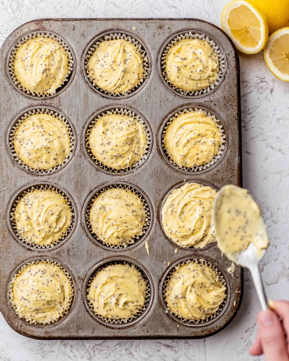 The image shows a metal muffin tray holding twelve cupcake liners filled with a light yellow batter speckled with small black poppy seeds. The batter looks creamy with a slightly rough texture and is roughly spread in each liner with swirled patterns on top. On the right side of the tray, a woman's hand is holding a spoon coated with the same poppy seed batter, and some batter drips from the spoon. The tray is set on a white marbled textured surface with a halved lemon placed near the top right corner as part of the background. photo taken with an iphone --ar 4:5 --v 7
