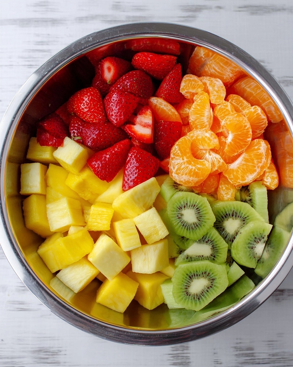 A shiny silver bowl sits on a white marbled surface filled with five distinct fruit layers arranged in sections. Starting from the top left, there are bright red quartered strawberries with visible seeds and smooth texture. Next to the strawberries to the right, vibrant orange mandarin segments with a slightly shiny surface. Below the mandarins, there are bright green kiwi slices showing their small black seeds and light green center. Below the kiwi, there are pale yellow pineapple chunks cut into small pieces with a slightly fibrous texture. Finally, at the bottom left, there are smooth, yellow mango cubes with a soft matte finish. The fruits are neatly separated inside the bowl, creating a colorful and fresh look. Photo taken with an iphone --ar 4:5 --v 7