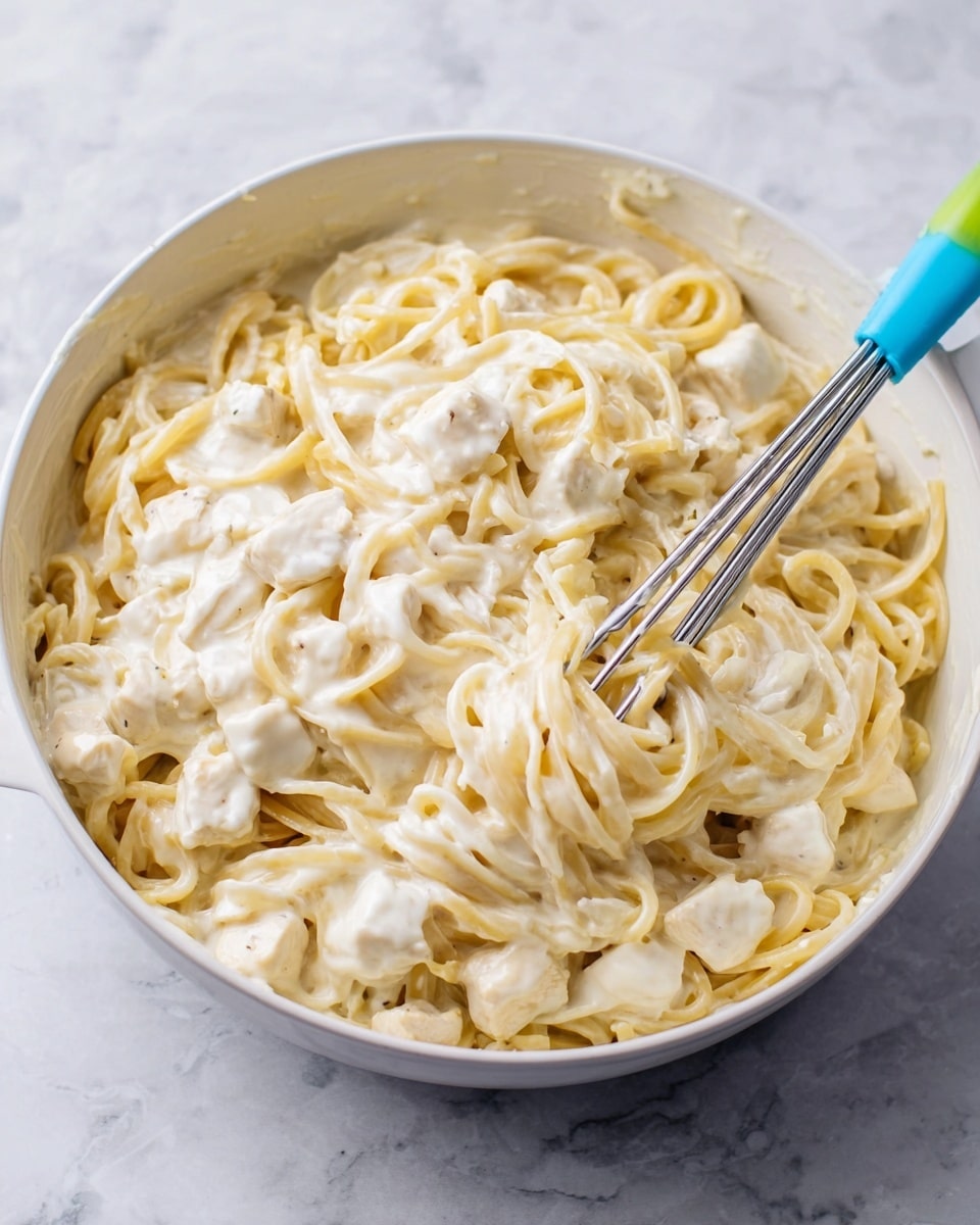 The image shows a white rectangular baking dish filled with a creamy pasta casserole. The top layer is golden melted cheese sprinkled with chopped green herbs. Beneath the cheese, there is a thick creamy sauce covering pieces of white meat and long pasta noodles. A metal serving spoon is scooping some of the casserole from the corner, revealing layers of pasta, sauce, and meat inside. The dish is placed on a white marbled surface with a soft striped cloth nearby. Photo taken with an iphone --ar 4:5 --v 7