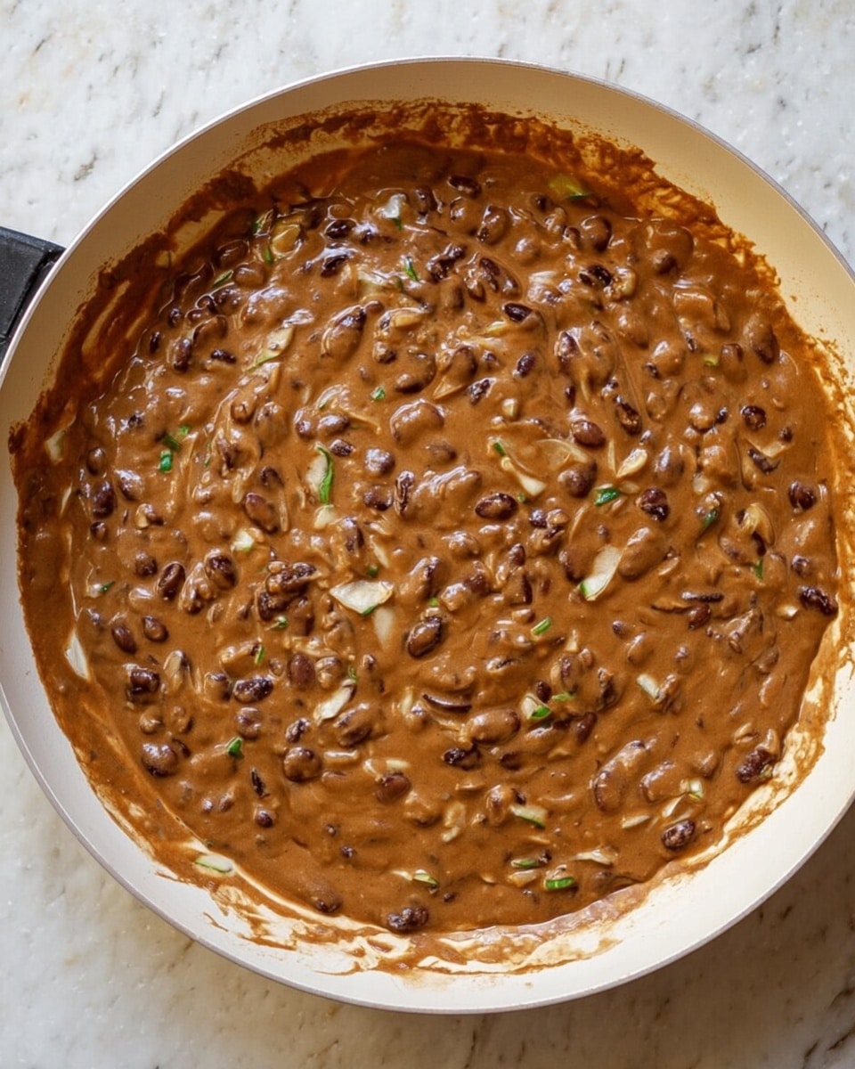 A creamy brown mixture of cooked black beans with some small green and white pieces, possibly vegetables, is shown in a white pan. The beans and sauce cover the entire bottom of the pan in one thick, uneven layer, with some sauce splashes around the pan edges. The texture looks soft and slightly chunky. The pan sits on a white marbled surface. photo taken with an iphone --ar 4:5 --v 7