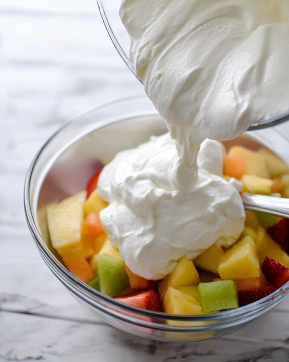 A close-up image shows a clear glass bowl pouring thick white whipped cream over a large silver bowl filled with colorful mixed fruit pieces. The fruit layer has chunks in yellow, orange, green, and red shades, looking fresh and juicy. A silver spoon rests inside the bowl among the fruit, partially visible under the cream. The scene is set on a white marbled surface, giving a clean and bright look. photo taken with an iphone --ar 4:5 --v 7