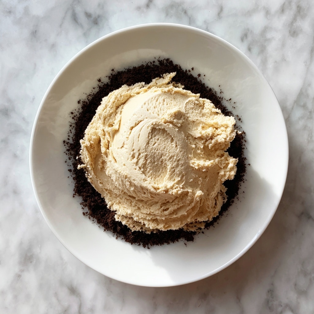 A white plate holds a single dark brown crumb layer spread evenly in a circle, forming the base. On top of the crumb base, there is a large, fluffy light tan cream layer placed roughly in the center with a soft, airy texture and uneven peaks and swirls. The background is a white marbled surface. Photo taken with an iphone --ar 4:5 --v 7