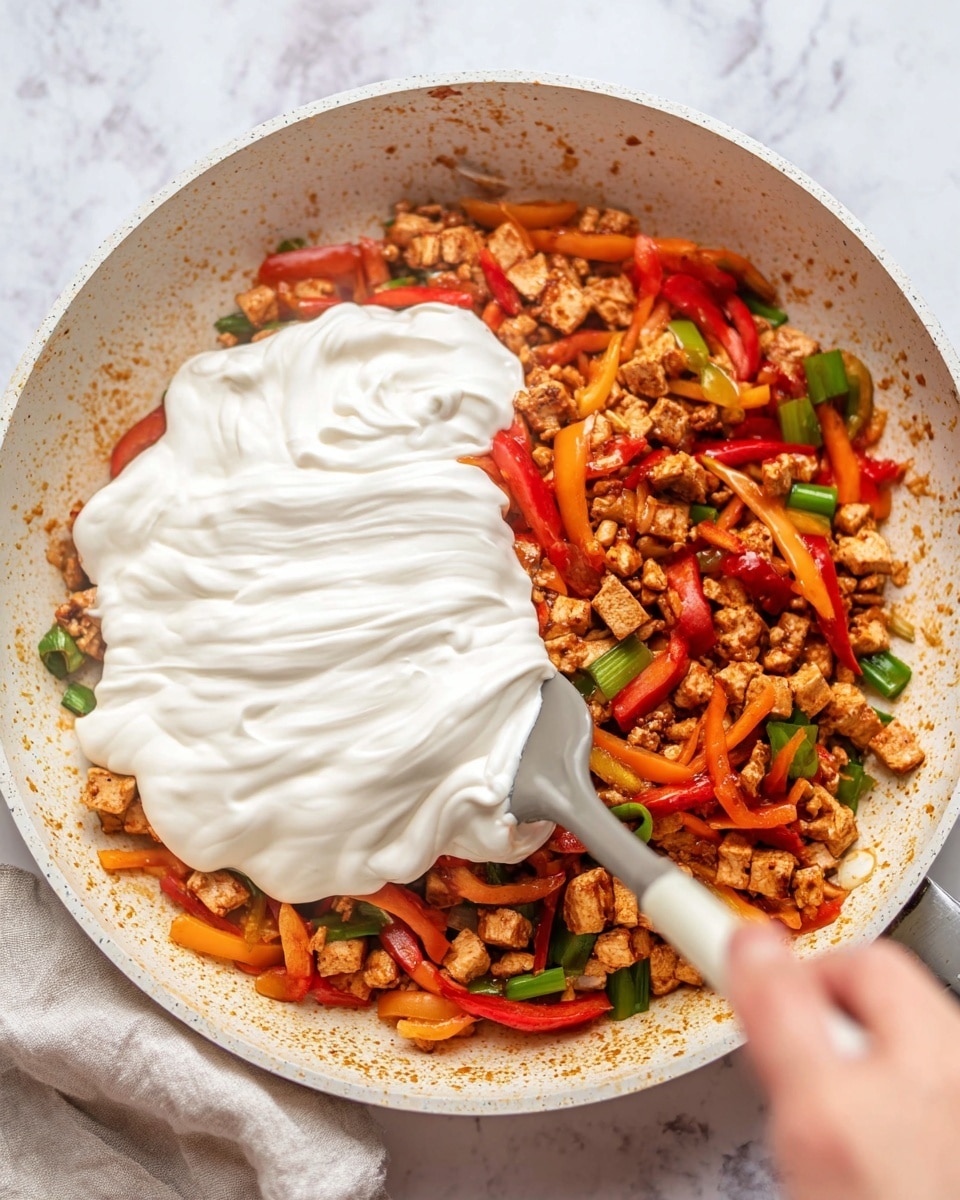 In a white frying pan on a white marbled surface, there is a mixed stir-fry with colorful layers. The bottom layer has small pieces of light brown tofu or meat, cooked with red and orange bell peppers cut into short strips, and green onion pieces scattered around. On top of this mixed stir-fry, a large spoon is adding a thick, white creamy layer smoothly spread over the food, creating a soft contrast to the vibrant vegetables and cooked pieces underneath. The edges of the pan show some spicy sauce stains. A woman's hand holds the spoon while spreading the cream. Photo taken with an iphone --ar 4:5 --v 7
