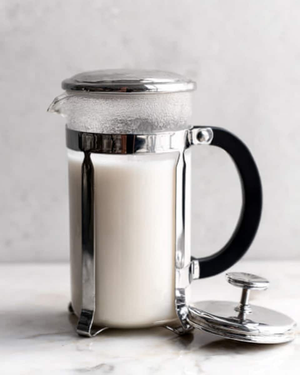 A clear glass French press filled with white milk sits on a white marbled surface. The French press has a shiny metal frame and a black handle on the right side. The lid with the plunger is placed next to the French press on the surface. The milk looks smooth and creamy inside the glass container photo taken with an iphone --ar 4:5 --v 7