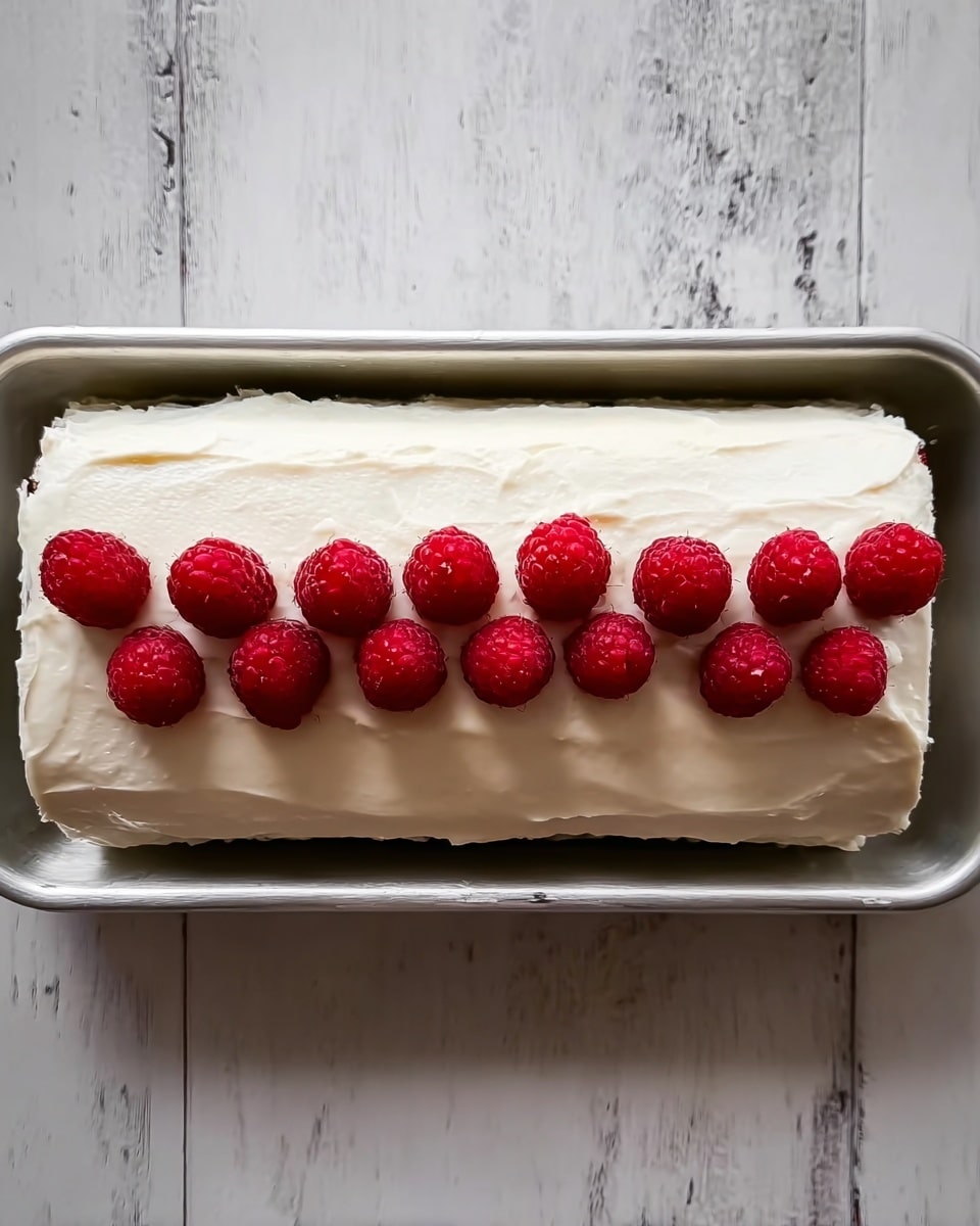A square sponge cake forms the base layer, pale yellow and soft in texture. On top is a smooth, even layer of red strawberry jam that reaches close to the edges. Above the jam is a thick, fluffy layer of white whipped cream, spread unevenly, creating soft peaks and waves. Scattered across the whipped cream are many bright red raspberries, spaced apart evenly. A woman's hand with a ring on the finger is gently pressing or adjusting one of the raspberries on the right side. The cake rests on a light fabric with thin dark stripes, set against a white marbled surface. Photo taken with an iphone --ar 4:5 --v 7