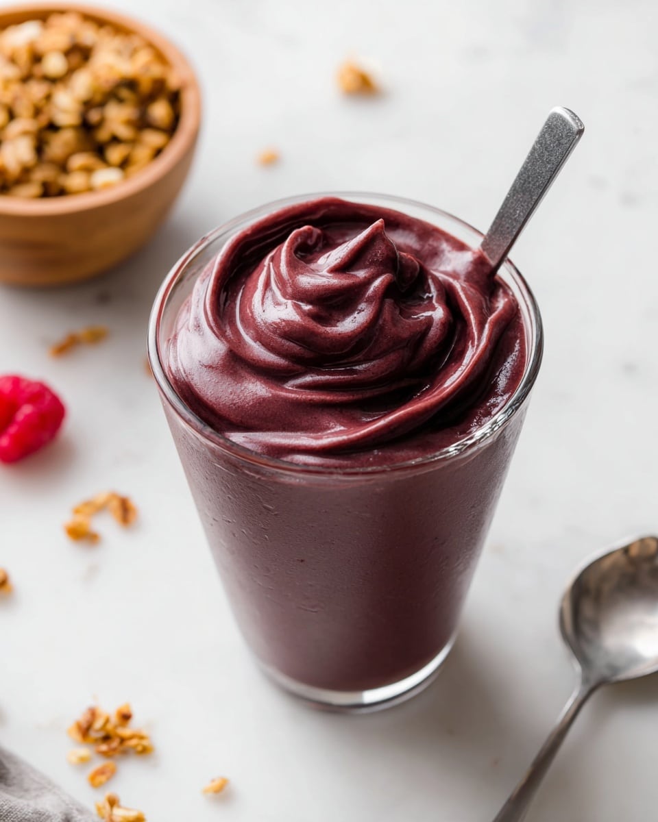 This image shows a thick, smooth dark purple smoothie served in a clear glass, filled to the brim with rich swirls and soft peaks on top. A metal spoon is placed inside the glass, partially immersed in the thick texture. In the blurred white marbled background, there is a wooden bowl filled with granola on the top left and a few scattered granola pieces and a raspberry nearby. A silver spoon handle is visible at the bottom right. The scene is bright and clean with a fresh and appetizing feel. photo taken with an iphone --ar 4:5 --v 7