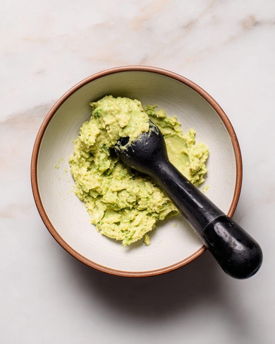 A white bowl with a brown rim sits on a white marbled surface. Inside the bowl is a small pile of light green mashed avocado with some texture from small lumps. Resting diagonally in the bowl is a black pestle with some avocado pasted on its rounded end. The scene is simple and clean, focused on the bowl and its contents. photo taken with an iphone --ar 4:5 --v 7