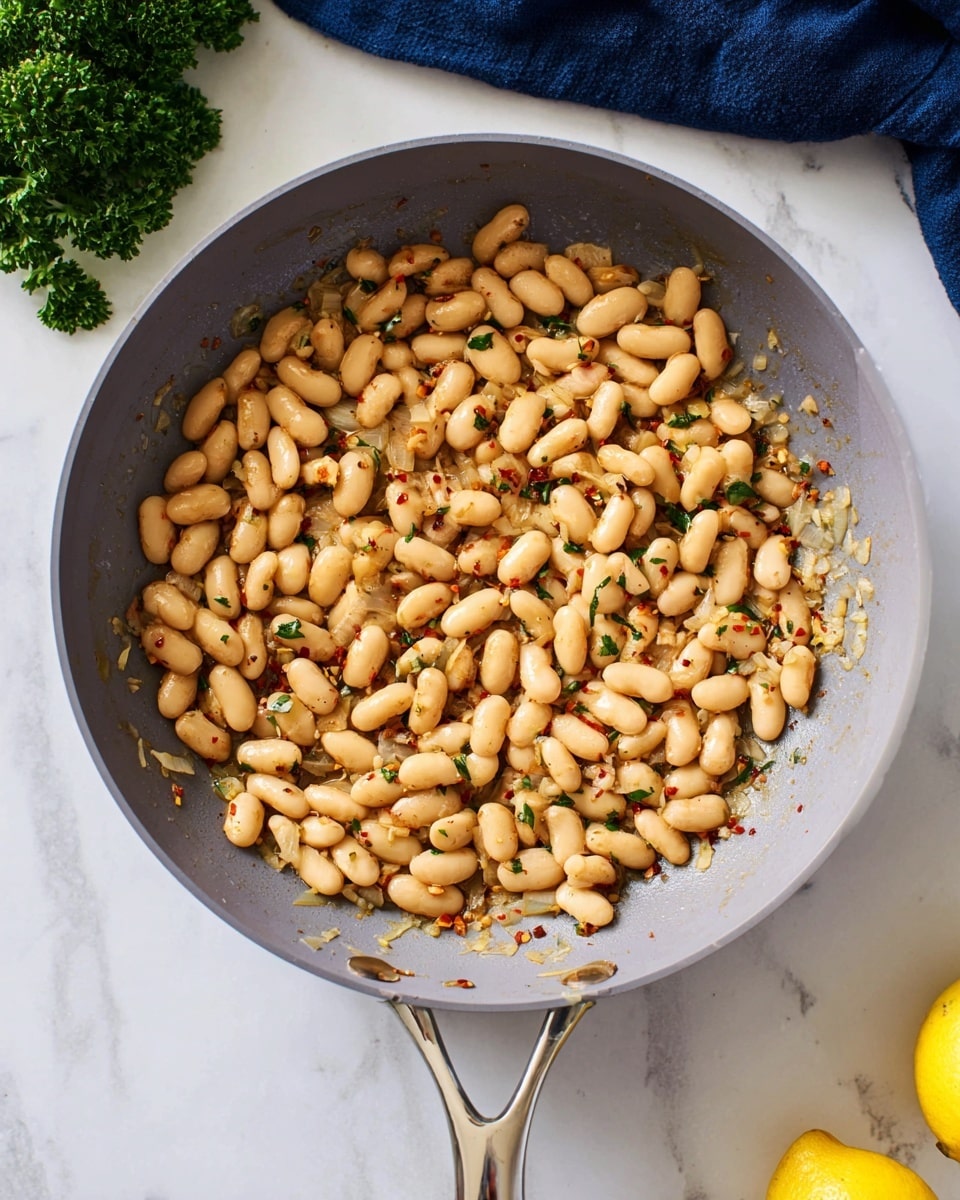 A light gray frying pan filled with cooked white beans, mixed with small bits of onions and red chili flakes, giving a speckled texture. The pan has a shiny metal handle. The pan is placed on a white marbled surface with a green bunch of parsley near the top edge and part of a lemon visible at the bottom right corner. A dark blue cloth is partially visible on the top left corner. photo taken with an iphone --ar 4:5 --v 7