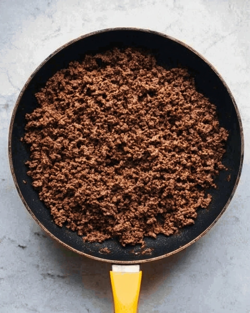The image shows a black frying pan with a yellow handle filled with cooked ground beef. The beef is evenly spread in one layer, with a rich brown color and a crumbly texture. The pan is placed on a white marbled surface. Photo taken with an iphone --ar 4:5 --v 7