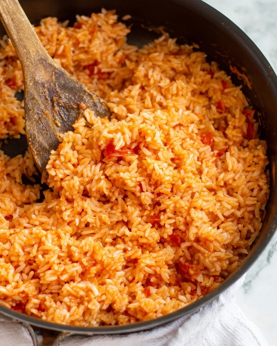 This image shows a close-up of a black pan filled with cooked rice mixed with tomato sauce, giving it an overall light orange color with red patches throughout. The rice grains look soft and fluffy, slightly sticky, with the tomato bits spread unevenly but visible. A wooden spoon is partly inside the pan, mixing the rice, with its textured surface showing some rice and sauce residue. The bottom edge of the image shows a white cloth. The background is a white marbled texture photo taken with an iphone --ar 4:5 --v 7