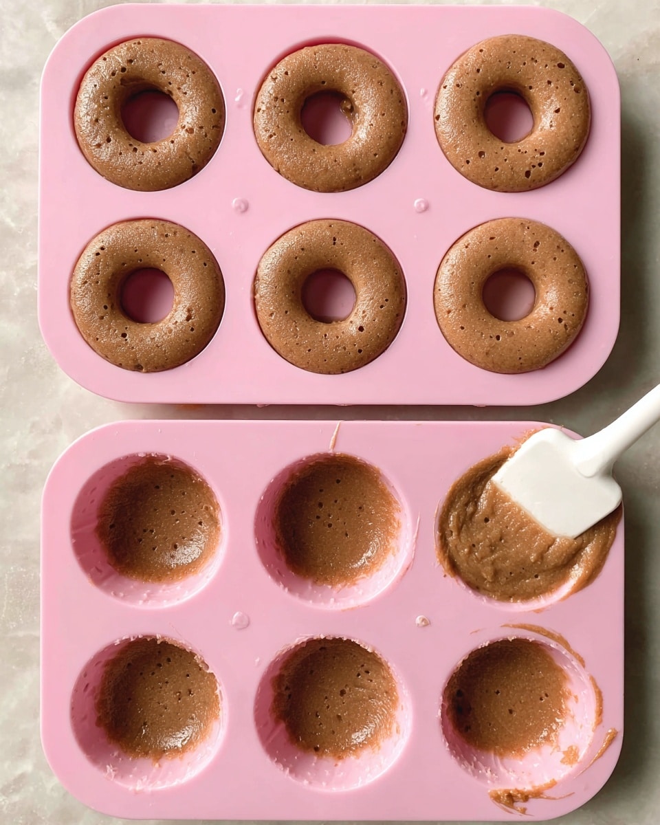 The image shows two pink silicone donut molds filled with brown dough. The top mold is fully filled with six round donut shapes, each having a smooth brown batter with specks on the surface. The bottom mold has four empty donut shapes on the left and two filled on the right, with a white spatula spreading the brown dough into one of the molds on the bottom right corner. The scene is set on a white marbled surface. Photo taken with an iphone --ar 4:5 --v 7