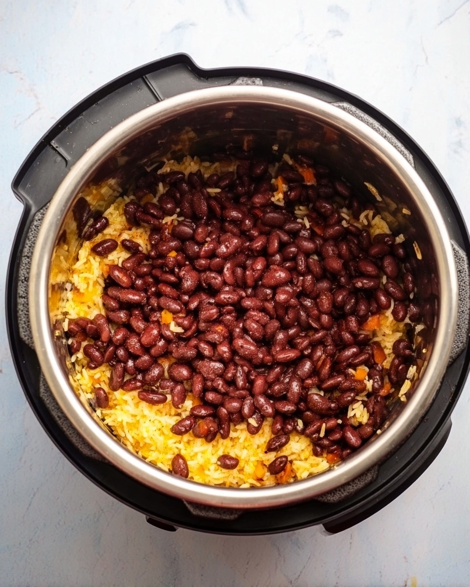 Inside a steel pot with a black outer base, there are two visible layers of food. The bottom layer is made of cooked rice that has a yellow-orange color with bits of small orange pieces mixed in. On top of this is a thick layer of dark red beans, fully covering the rice. The pot is placed on a white marbled surface and the photo is taken from above. photo taken with an iphone --ar 4:5 --v 7