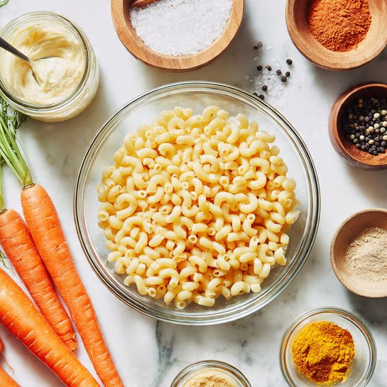 A clear glass bowl in the center filled with uncooked elbow macaroni pasta in a light yellow color. Around the bowl, on a white marbled surface, there are bright orange carrots with green tops on the left side. To the top left, there is an open jar of mayonnaise with a small spoon next to it. Above the pasta bowl, a small wooden bowl holds white salt, to its right a small glass bowl with reddish-orange powder, next to it a small glass jar with a beige powder, and further right, a black pepper grinder with black peppercorns scattered nearby. Below the pasta bowl, a small jar of bright yellow turmeric powder and, further down, two small glass jars with fine powders are visible. On the right side near the bottom, a small glass bowl holds white salt with a silver tablespoon resting in it. photo taken with an iphone --ar 4:5 --v 7