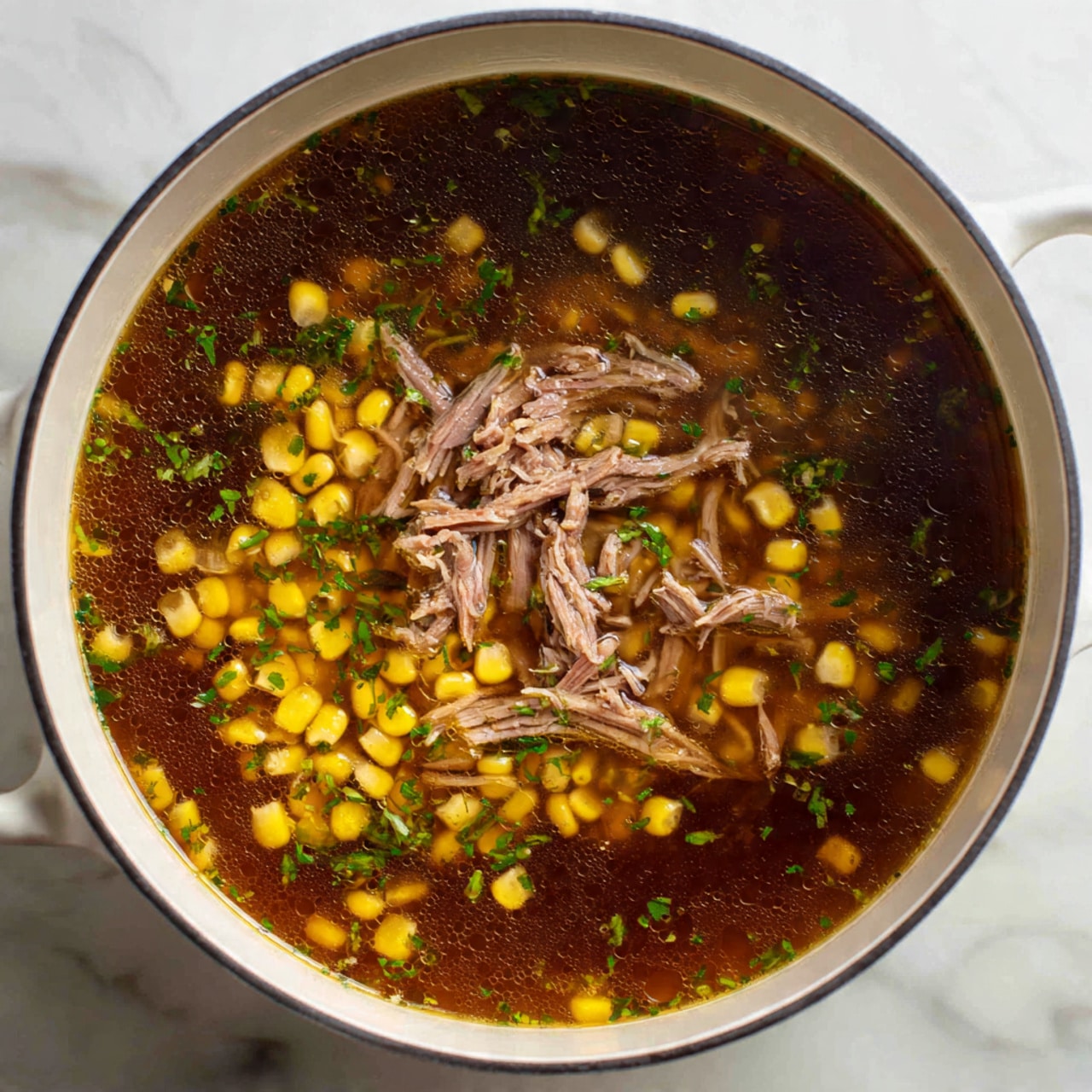 The image shows a close-up view of a pot filled with a dark brown broth containing a large amount of yellow corn kernels and green herbs floating around. On top of the broth and corn, there is a generous layer of light pink shredded meat pieces, slightly moist and fibrous in texture. The pot's inner surface is white, and the photo is taken on a white marbled surface. photo taken with an iphone --ar 4:5 --v 7