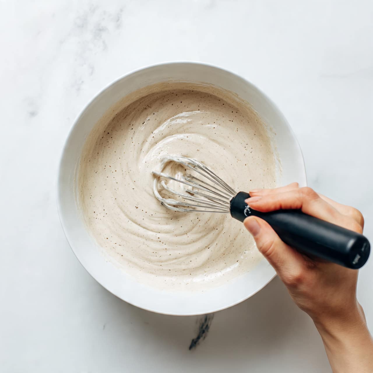 A white bowl filled with a thick, light beige foam being stirred by a woman's hand holding a small black electric whisk. The foam has a smooth texture with some bubbles on the surface. The bowl is placed on a white marbled surface, and the woman's hand enters the frame from the right side. Photo taken with an iphone --ar 4:5 --v 7