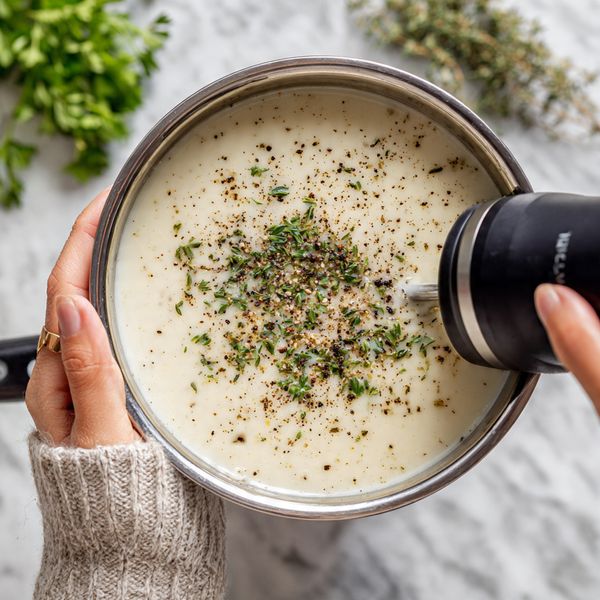A close-up of a silver pot filled with creamy white soup that has small green pieces and black pepper sprinkled on top. A woman's hand holds a black immersion blender, blending the soup inside the pot. The background surface is a white marbled texture with some green herbs blurred in the background. photo taken with an iphone --ar 4:5 --v 7