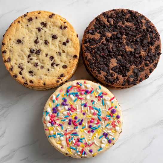 Three large round cookie cakes lie flat on a white marbled surface. The top left cookie is a classic chocolate chip cookie with a golden brown edge and light tan center speckled with dark chocolate chips. The top right cookie is a deep dark brown chocolate cookie cake studded with similarly dark chocolate chips, its texture smooth and slightly cracked. The bottom cookie is pale with white chocolate chips and brightly colored sprinkles in red, blue, yellow, purple, and pink spread all over its surface. Each cookie cake has a slightly raised edge with a smooth, even baked texture. Photo taken with an iphone --ar 4:5 --v 7