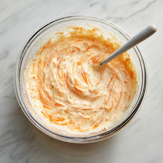 A clear glass bowl filled with a creamy, thick mixture that is swirled with light and darker orange colors, showing a rich, smooth texture with small bits throughout. A light grey spoon is partially placed in the bowl, mixing the creamy content. The bowl sits on a white marbled surface. photo taken with an iphone --ar 4:5 --v 7