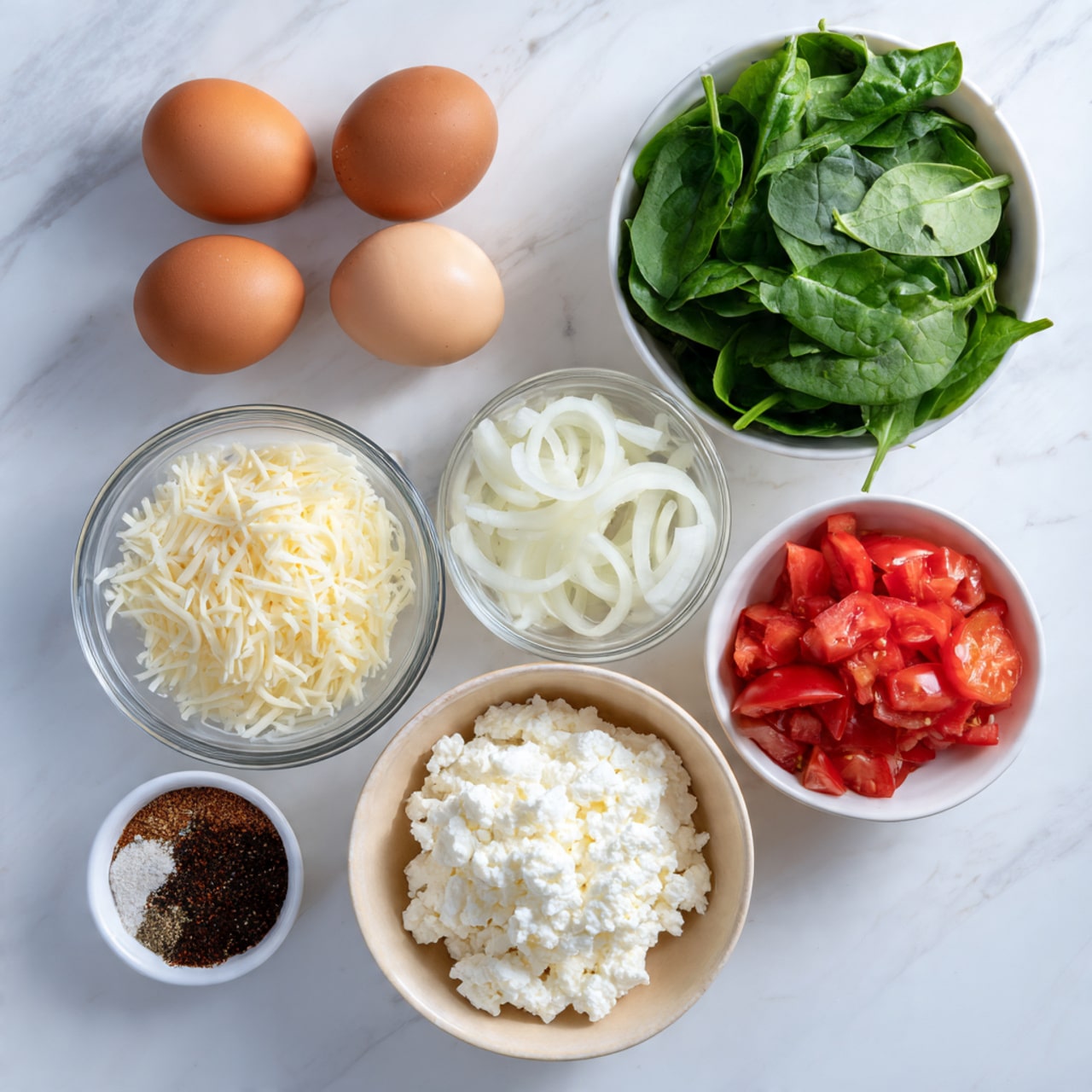 The image shows several cooking ingredients on a white marbled surface. There are four brown eggs arranged loosely on the top left. Below the eggs are two small clear glass bowls: one with white shredded cheese and the other with white sliced onions. To the right, there is a white bowl filled with fresh green spinach leaves and another white bowl with chopped red tomatoes. At the bottom left, there is a small brown bowl containing light brown and black ground spices. In the center bottom, there is a larger beige bowl filled with white cottage cheese. photo taken with an iphone --ar 4:5 --v 7