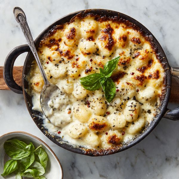 This image shows a close-up of a black cast iron skillet filled with a creamy cauliflower baked dish. The top layer is golden and slightly browned melted cheese with red pepper flakes sprinkled across. Under the cheese, a thick, white, creamy sauce covers cauliflower florets, which are dense and soft. A spoon lifts a portion from the skillet, showing sticky, stretchy melted cheese strings falling back into the skillet. Bright green basil leaves sit on the right side, adding a fresh pop of color. The background features a white marbled surface and a blurred amber-colored glass. Photo taken with an iphone --ar 4:5 --v 7