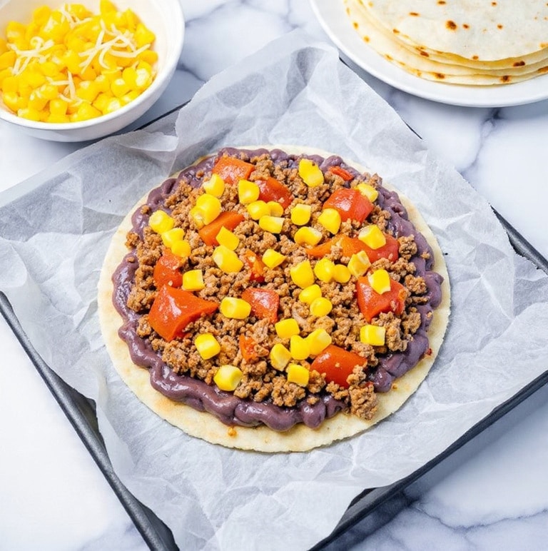 A round tortilla with three layers is shown on a white marbled surface lined with parchment paper on a baking tray. The base layer is a purple-black bean spread that covers the tortilla fully. On top of that is a layer of cooked ground beef with a brown, crumbly texture mixed with small, vibrant red tomato chunks. The top layer is scattered yellow corn kernels, adding bright pops of color. Nearby, there are small white bowls filled with corn and shredded cheese, and a stack of tortillas on a white plate partially in view. photo taken with an iphone --ar 4:5 --v 7