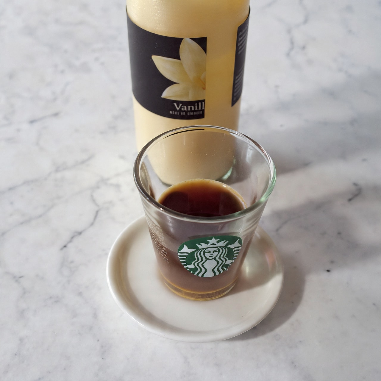 A clear glass cup with a small amount of dark brown liquid at the bottom, showing the Starbucks logo in green on the side, sits on a round white coaster. Behind the cup, there is a tall clear bottle with a yellow and black label that says