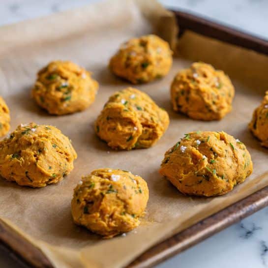 The image shows multiple small rounded mounds of a mixed orange and white textured dough placed on brown parchment paper. Each mound has a rough, uneven surface with visible small chunks and flecks of herbs evenly spread throughout. They are spaced evenly apart in a grid pattern, resting on a flat baking tray. The lighting is warm and soft, highlighting the texture and the slight shine of the mixture. The background surface is a white marbled texture photo taken with an iphone --ar 4:5 --v 7