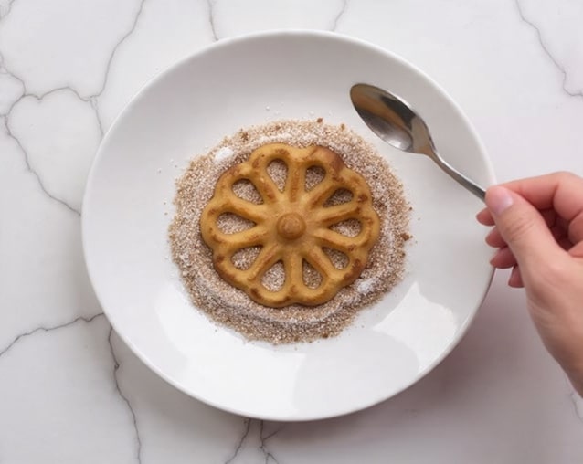 A light brown flower-shaped cookie with eight petal-like openings and a small round center sits in the middle of a shallow white plate. Around and under the cookie is a mix of white and light brown granulated sugar, covering most of the plate’s bottom in a rough circle. A woman's hand holding a silver spoon is visible on the right side, slightly above the plate. The plate is placed on a white marbled surface. Photo taken with an iphone --ar 4:5 --v 7