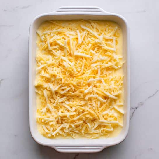 A white rectangular baking dish is filled with a single layer of smooth, creamy light yellow mixture topped evenly with shredded cheese that is a mix of pale yellow and white strands. The texture of the cheese is slightly stringy and loose, scattered to almost cover the entire surface, with some small gaps showing the creamy base underneath. The dish is set on a white marbled surface. photo taken with an iphone --ar 4:5 --v 7