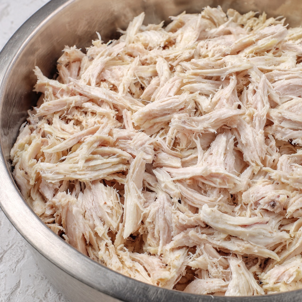 The image shows shredded cooked white meat piled in a large round metal bowl. The shredded pieces vary in size and thickness, with a fibrous texture and slight pinkish-white color. The meat strands are loosely arranged, filling almost the whole bowl. The background is a white marbled texture. Photo taken with an iphone --ar 4:5 --v 7