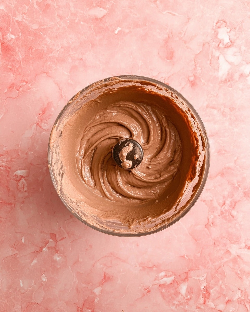 A top view of a round blender bowl filled with smooth, light brown chocolate mixture. The mixture has a creamy texture, with a swirl pattern around the center where a blender blade is visible. The blender blade is covered partly with the chocolate mix. The background is a white marbled texture but appears pink in this instance. photo taken with an iphone --ar 4:5 --v 7
