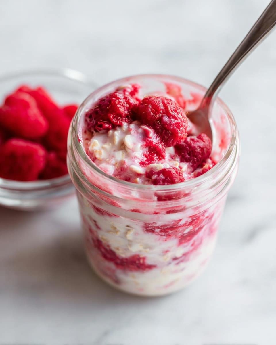 The image shows a clear glass jar filled with a mixed dish of creamy white oats and vibrant red raspberries. There are visible layers where the raspberries mix into the oats, creating swirls of deep pink and red throughout the creamy white base. Several whole raspberries sit mixed inside the oats, giving the texture a rough, chunky look. A metal spoon rests inside the jar, partially submerged in the mixture. The jar is placed on a white marbled surface, and there is a small clear bowl with fresh red raspberries in the background. Photo taken with an iphone --ar 4:5 --v 7