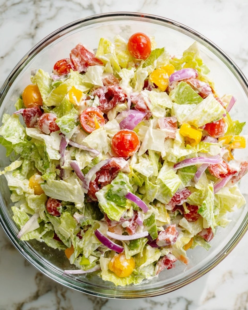 A clear glass bowl holds a colorful salad with many layers. The base layer is made of chopped light green and pale yellow lettuce leaves, mixed together with thin slices of white and purple onion. On top, there are bright red cherry tomato halves scattered evenly around. Small pieces of pale yellow cheese and slices of yellow pepper add more color. The salad is coated with a creamy white dressing that gives a shiny texture to the whole dish. The bowl sits on a white marbled surface. photo taken with an iphone --ar 4:5 --v 7