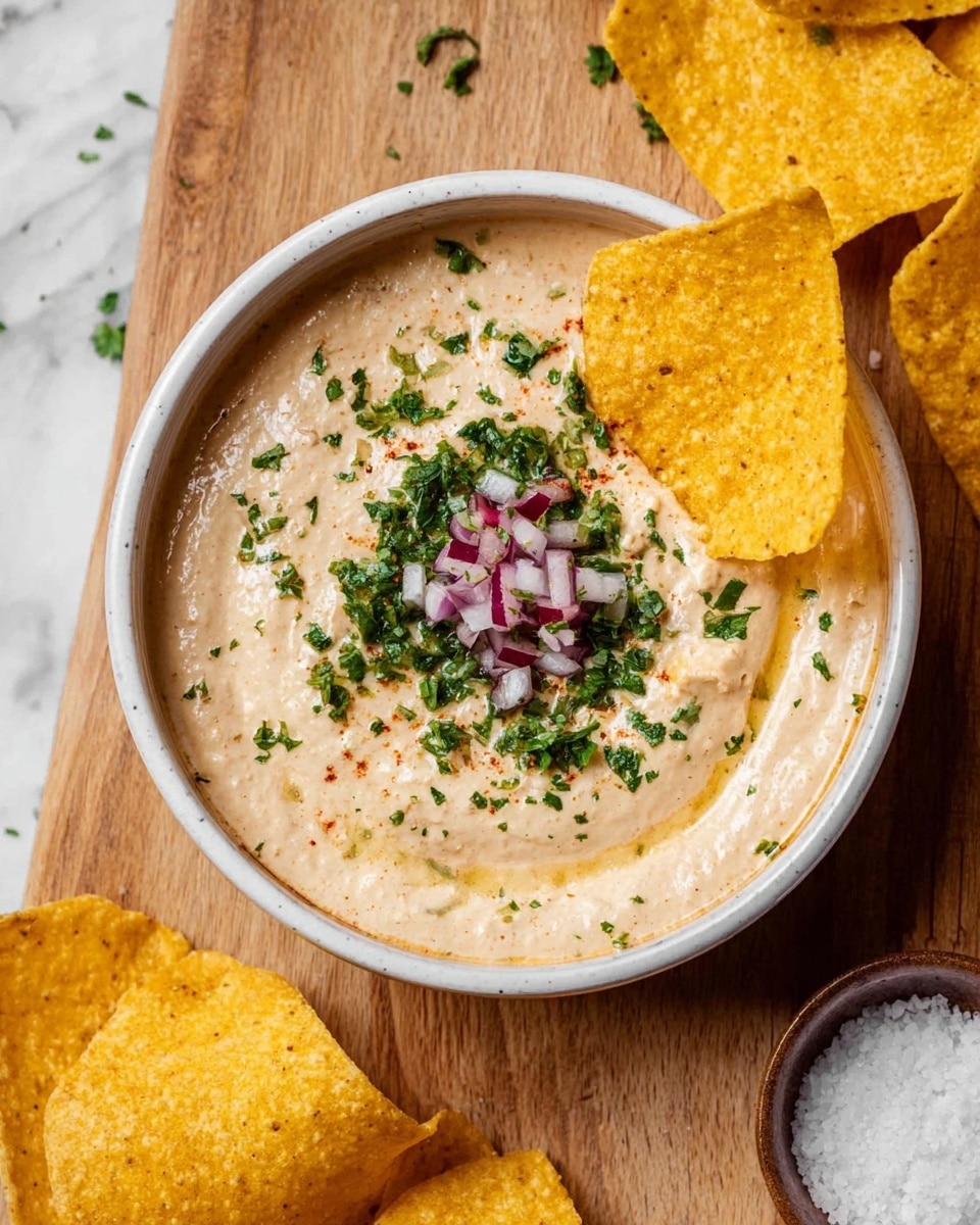 A white bowl filled with smooth, creamy beige dip sprinkled with finely chopped green herbs and small pieces of diced red onion in the center; two golden yellow tortilla chips are partially dipped into the right side of the bowl, while more yellow chips rest around the bowl on a wooden surface with a white marbled background. A small dish with coarse salt is partially visible at the bottom right corner. Photo taken with an iphone --ar 4:5 --v 7