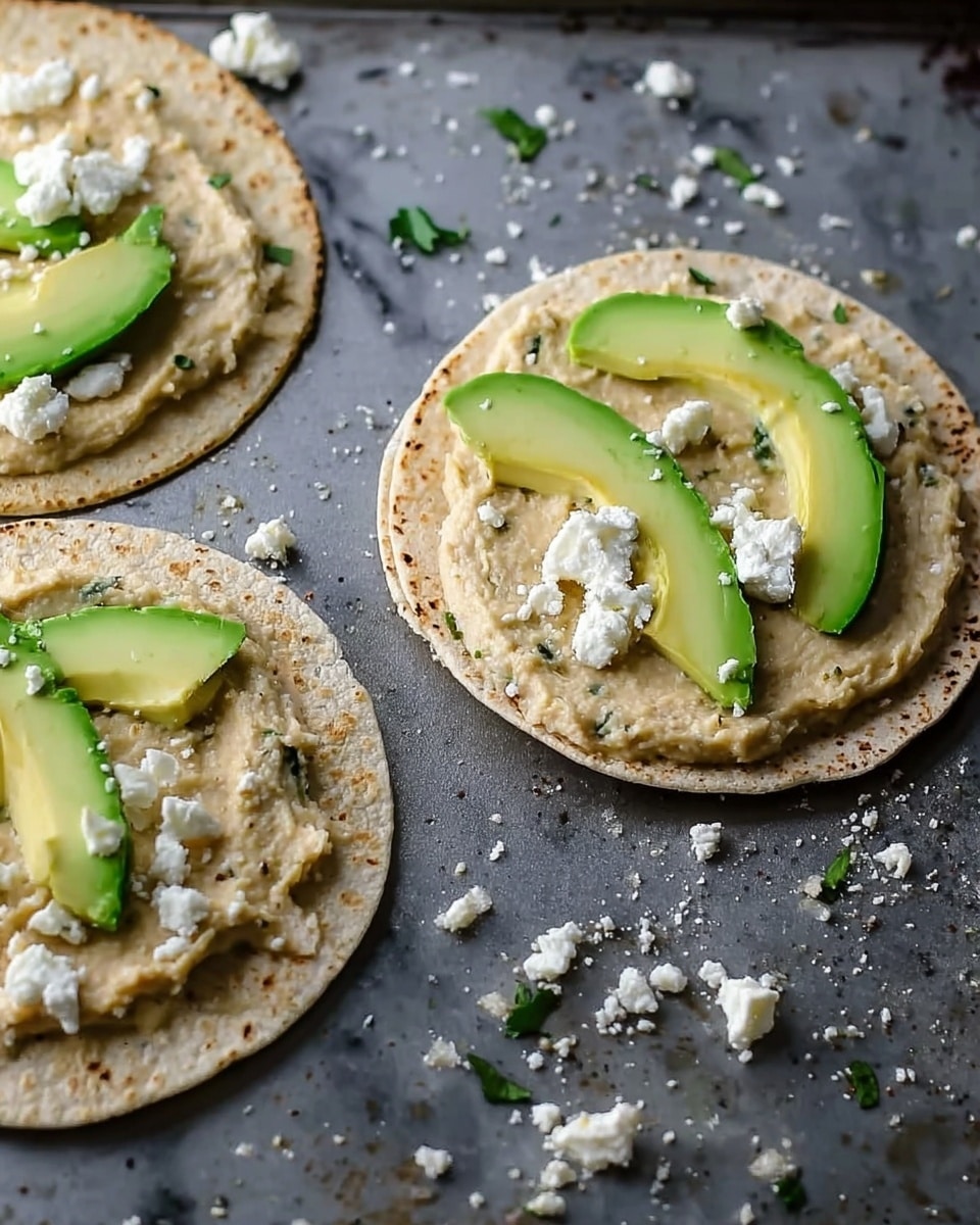Three small round tortillas lie on a dark baking tray, each topped with a beige spread that has green specks in it. On two tortillas, smooth pale green slices of avocado are placed near the center. White crumbly cheese is scattered on top of the avocado on one tortilla and sprinkled around the tray. The tortillas have a soft texture and look fresh. The background is a white marbled surface. Photo taken with an iphone --ar 4:5 --v 7