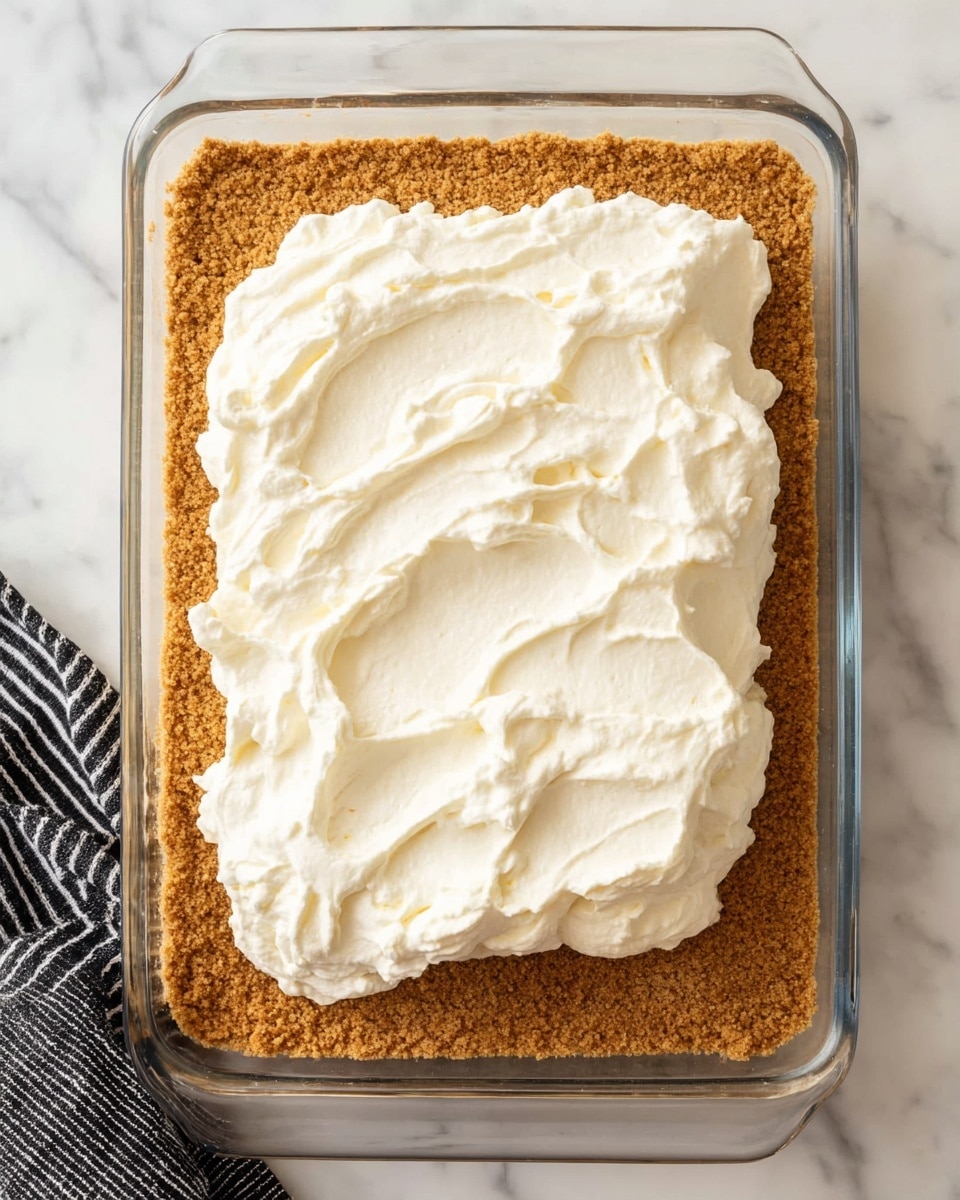 A clear rectangular glass dish is filled with a thick, even layer of light brown crumb crust that covers the bottom fully. On top of this crust, near the center, there is a large, fluffy, uneven mound of smooth, white cream spread loosely with soft peaks and swirls, creating a contrast in color and texture between the crumb layer and the cream. The dish is placed on a white marbled surface, and the corner of a black and white striped cloth is visible at the bottom left edge of the image. photo taken with an iphone --ar 4:5 --v 7
