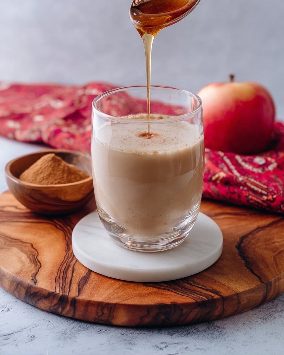 A clear glass filled with a beige creamy liquid sits on a round white marble coaster placed on a wooden board with rich brown swirls; a spoon above the glass pours a thin stream of amber liquid into it. Behind the glass, there is a small wooden bowl filled with brown powder, a red apple, and a red patterned cloth resting on a white marbled surface. photo taken with an iphone --ar 4:5 --v 7