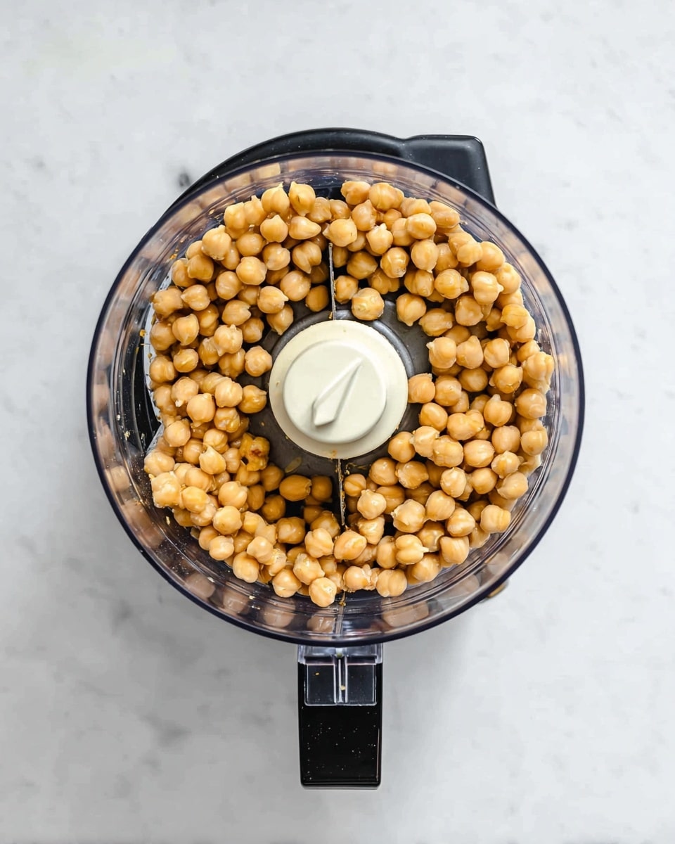 The image shows a black food processor on a white marbled surface from above. Inside the clear plastic bowl of the food processor, a single layer of light brown chickpeas fills the space around the sharp white blade in the center. The chickpeas are evenly spread, covering most of the bowl but leaving the blade visible. The photo taken with an iphone --ar 4:5 --v 7