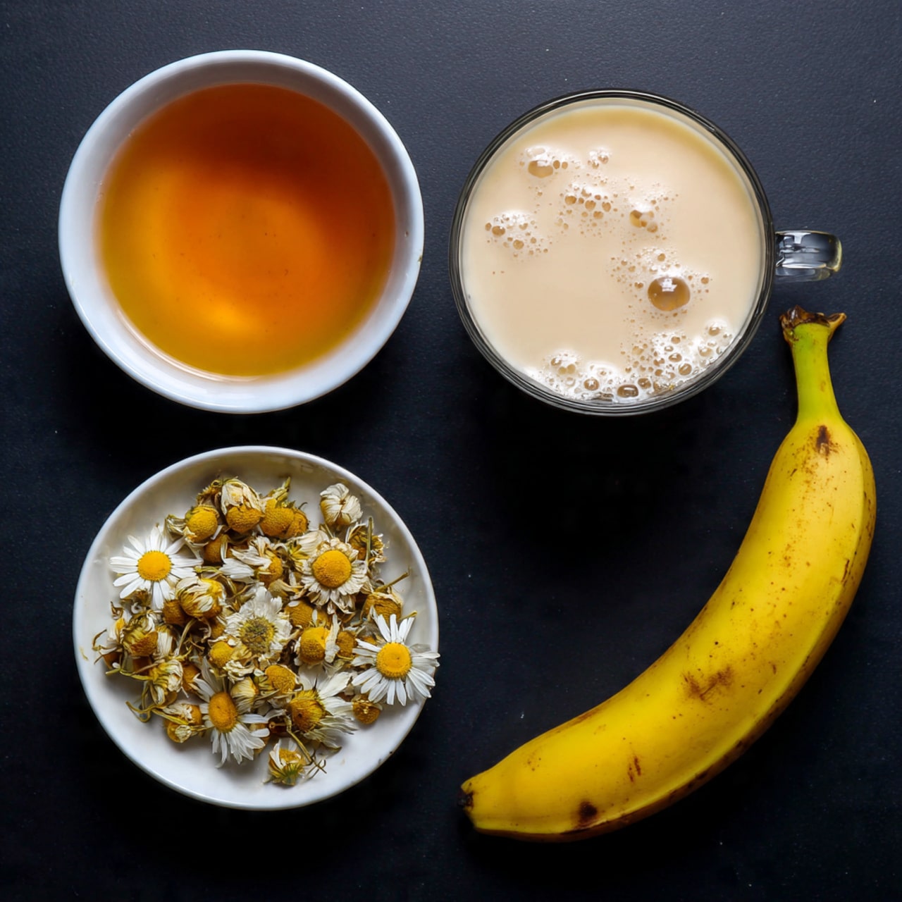 The image shows four items placed on a black surface: at the top left is a white bowl filled with a golden amber liquid, at the top right is a clear glass mug filled with creamy beige liquid with bubbles on the surface, at the bottom left is a white bowl containing dried chamomile flowers with yellow centers and pale petals, and at the bottom right is a whole ripe yellow banana with a few dark spots on the peel. photo taken with an iphone --ar 4:5 --v 7