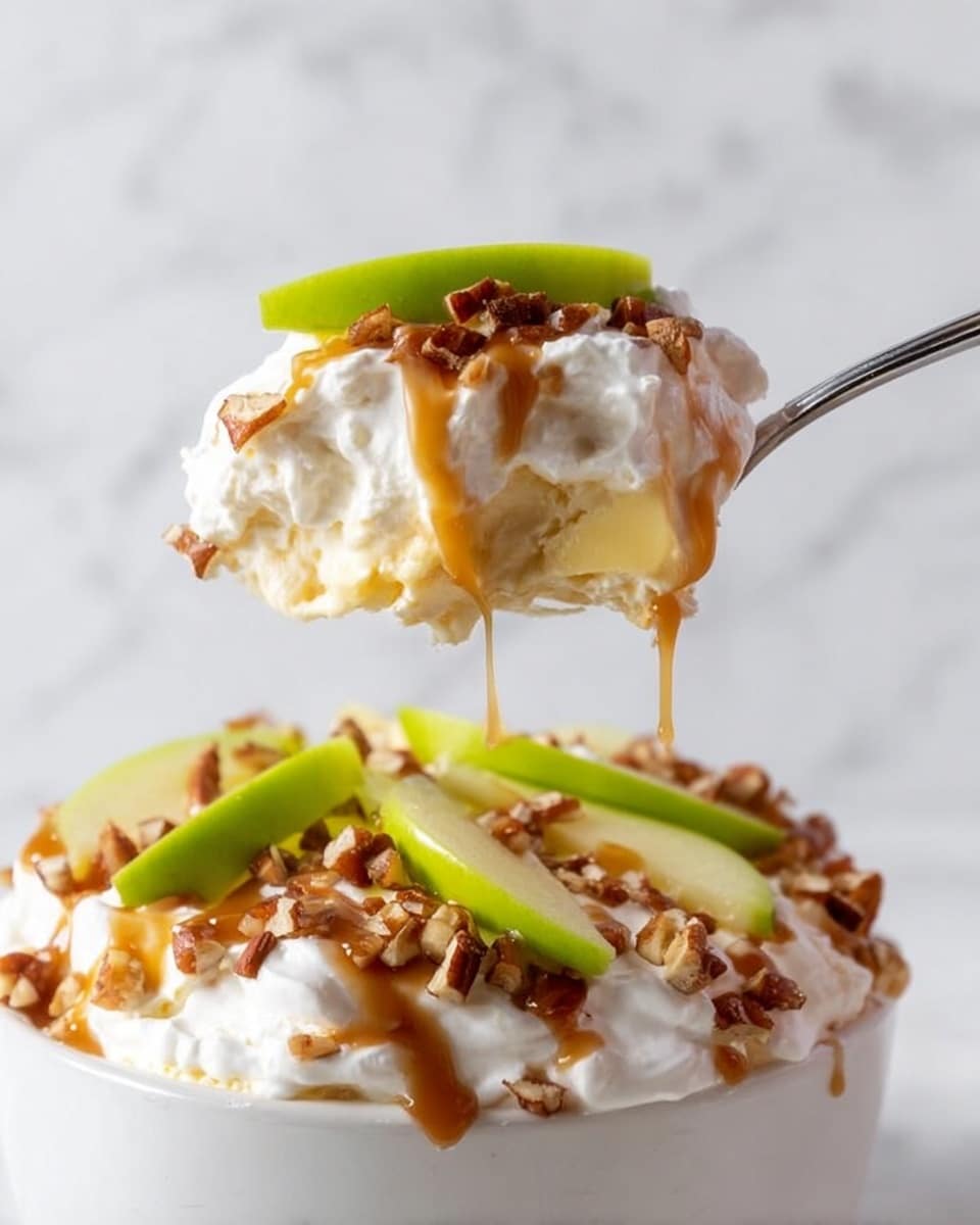 A spoon holds a layered dessert showing creamy white whipped cream at the bottom, topped with a light yellow layer of smooth filling, drizzled with caramel sauce, and sprinkled with small brown nut pieces. On top of the spoon’s dessert, there is a thin slice of bright green apple. Below the spoon, the white bowl is filled with the same layered dessert, topped with more whipped cream, caramel drizzle, chopped nuts, and green apple slices scattered on top. The background is a white marbled texture. photo taken with an iphone --ar 4:5 --v 7