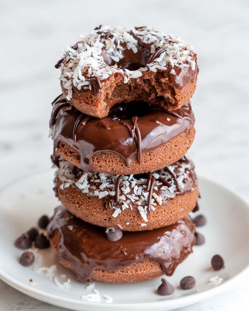 A stack of four chocolate donuts sits on a white plate against a white marbled background. The bottom donut is thick, with a light brown color and topped with chocolate glaze, small chocolate chips, and white shredded coconut flakes. The next donut up is covered with smooth chocolate glaze and decorated with thin dark chocolate sprinkles. Above that is another donut with glossy chocolate glaze dripping down the sides and small chocolate chips. The top donut has a bite taken out of it, showing a soft, moist inside and is topped with a layer of chocolate glaze and white shredded coconut flakes scattered on top. Photo taken with an iphone --ar 4:5 --v 7