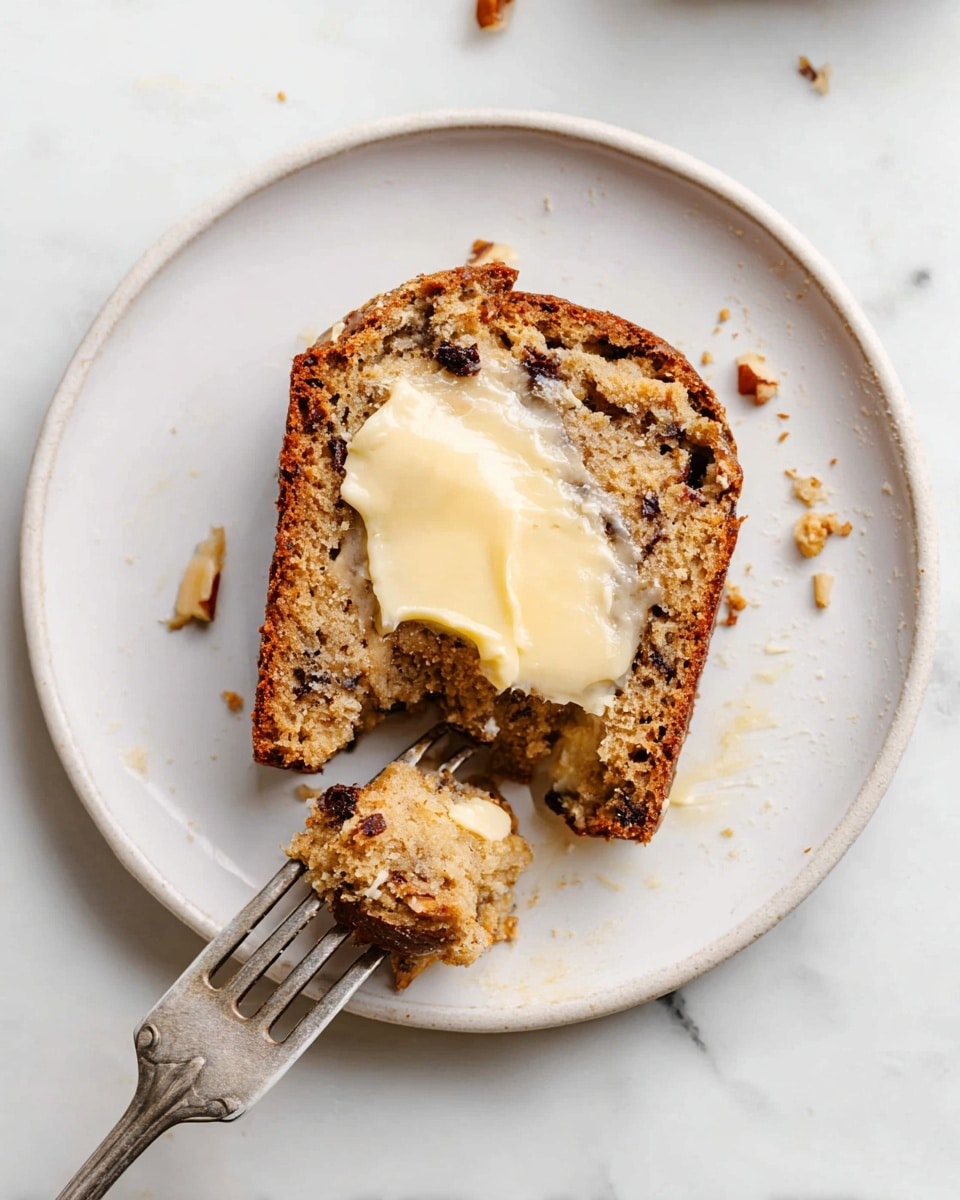 A single slice of light brown banana bread with visible darker spots of nuts and chocolate chips sits in the center of a white plate. The top of the bread is topped with a melting creamy light yellow spread, partially dripping down the sides. A silver fork rests on the plate, with its prongs inserted into a small bitten piece of the bread near the bottom left. Small crumbs and pieces of nuts are scattered around the plate and on the white marbled surface below. Photo taken with an iphone --ar 4:5 --v 7