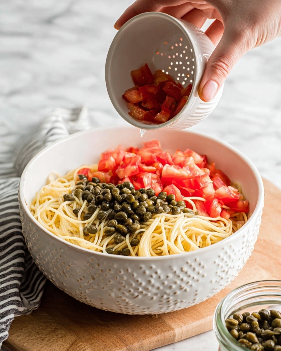 A white bowl sits on a white marbled surface filled halfway with thin, pale yellow spaghetti. On top, one side is covered with small, dark green capers piled in a heap, and the other side shows many small red tomato chunks. Above the bowl, a woman's hand tilts a small white bowl with raised dots on the inner surface, pouring more red tomato pieces into the larger bowl. A partly open jar with dark green capers is on the bottom right near the bowl. photo taken with an iphone --ar 4:5 --v 7