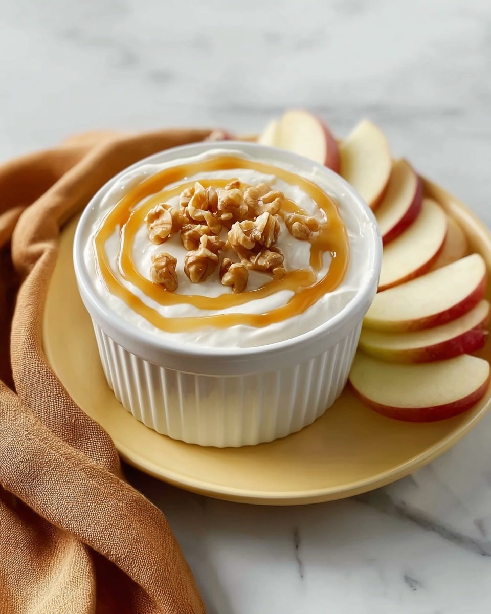 A small white ceramic ramekin filled with a creamy white dip topped with chopped nuts scattered across the surface. A red apple slice is dipped into the dip, catching golden caramel sauce that is being poured down, dripping off the apple slice and pooling on the dip. Several more red apple slices rest on a white plate below the ramekin. The background and surface feature a white marbled texture. Photo taken with an iphone --ar 4:5 --v 7